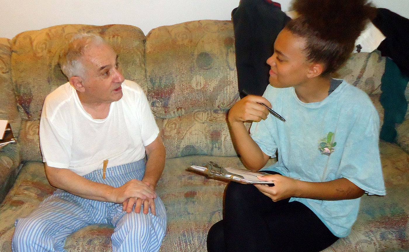 A young woman with a clipboard and pen interestedly looking at a grandparent who is answering family history questions.